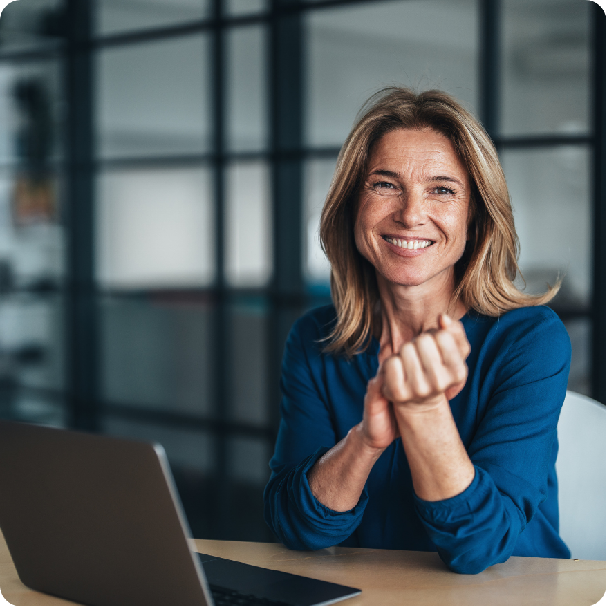 Smiling professional woman sitting at a desk with a laptop, confidently facing the camera in a modern office setting.